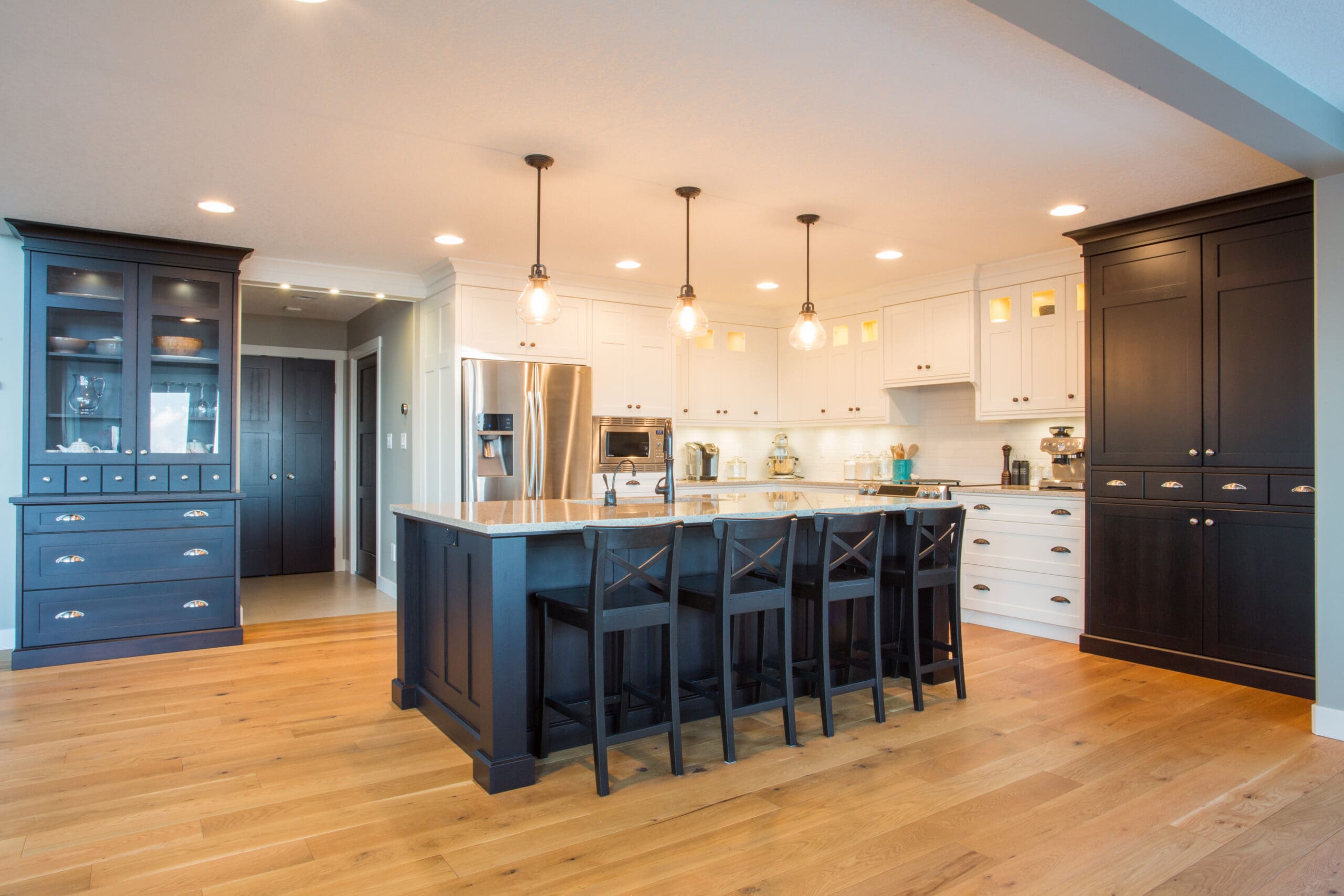 Modern kitchen with wood floors, a large island with four black chairs, pendant lights, stainless steel appliances, and white and dark cabinets—beautifully maintained by a property maintenance company. The room is brightly lit and spacious.
