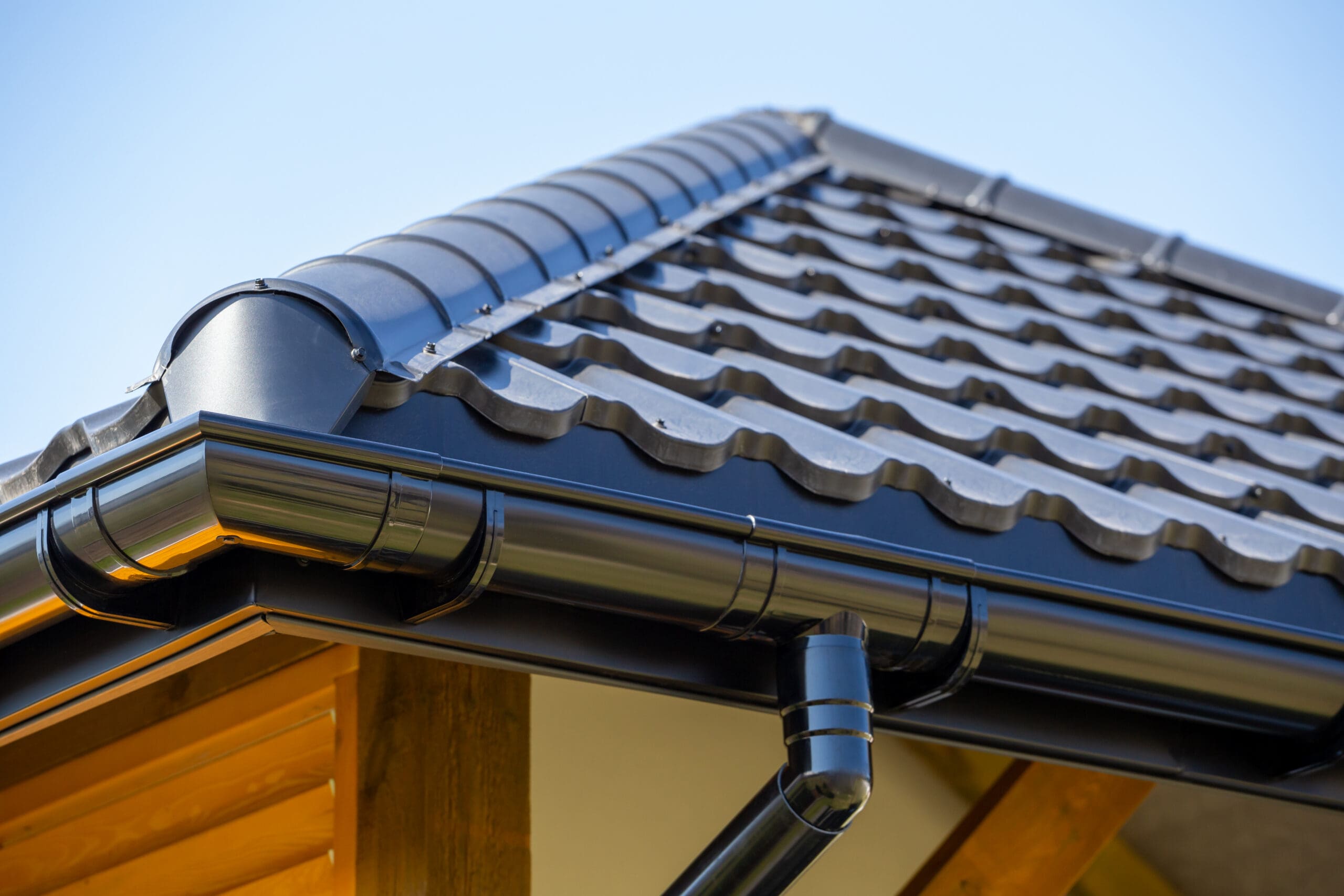 Close-up of a house roof with dark gray, wavy metal tiles and a matching black rain gutter system, showcasing expert fascia and guttering installation. The house exterior features wooden elements beneath a clear blue sky.
