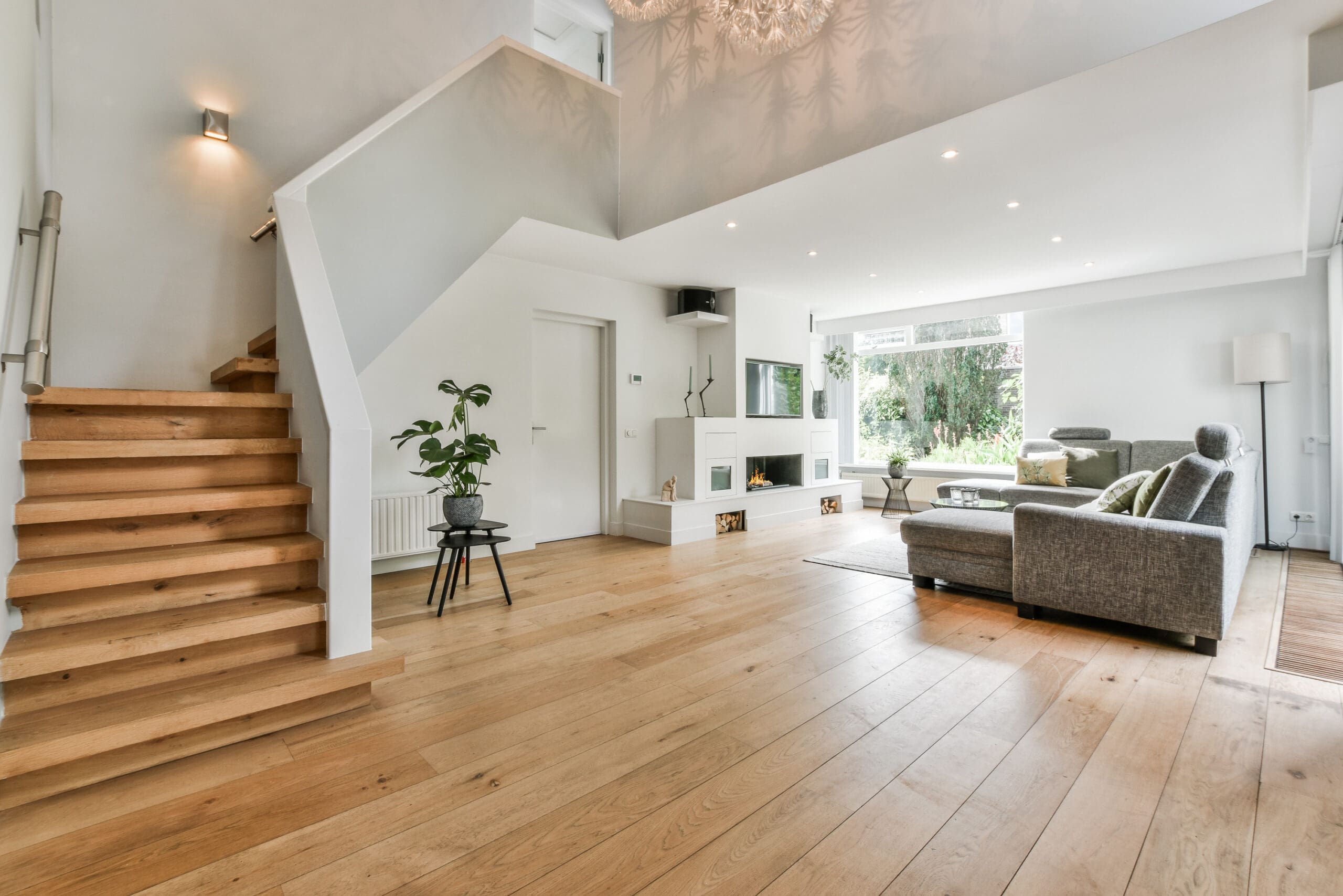 Modern living room with light wooden floors, gray sectional sofa, large windows, a plant on a stool, and open staircase with wooden steps; bright and minimalistic decor—perfect for those interested in stylish decorating.