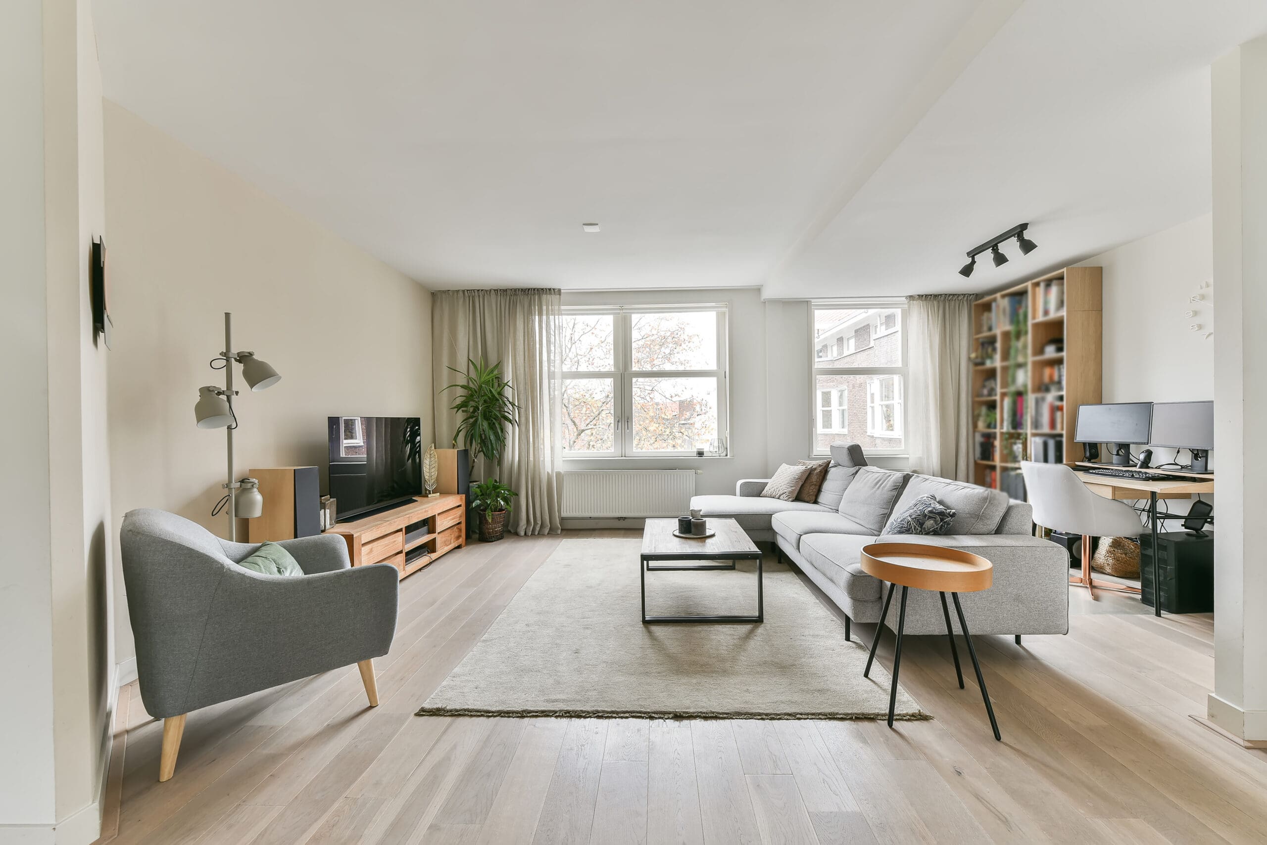 Modern living room with light wood floors, gray sofa, armchair, coffee table, and TV. Large windows bring in natural light. Flooring installation highlights the cozy, minimalist space with a desk and bookshelves in the corner.