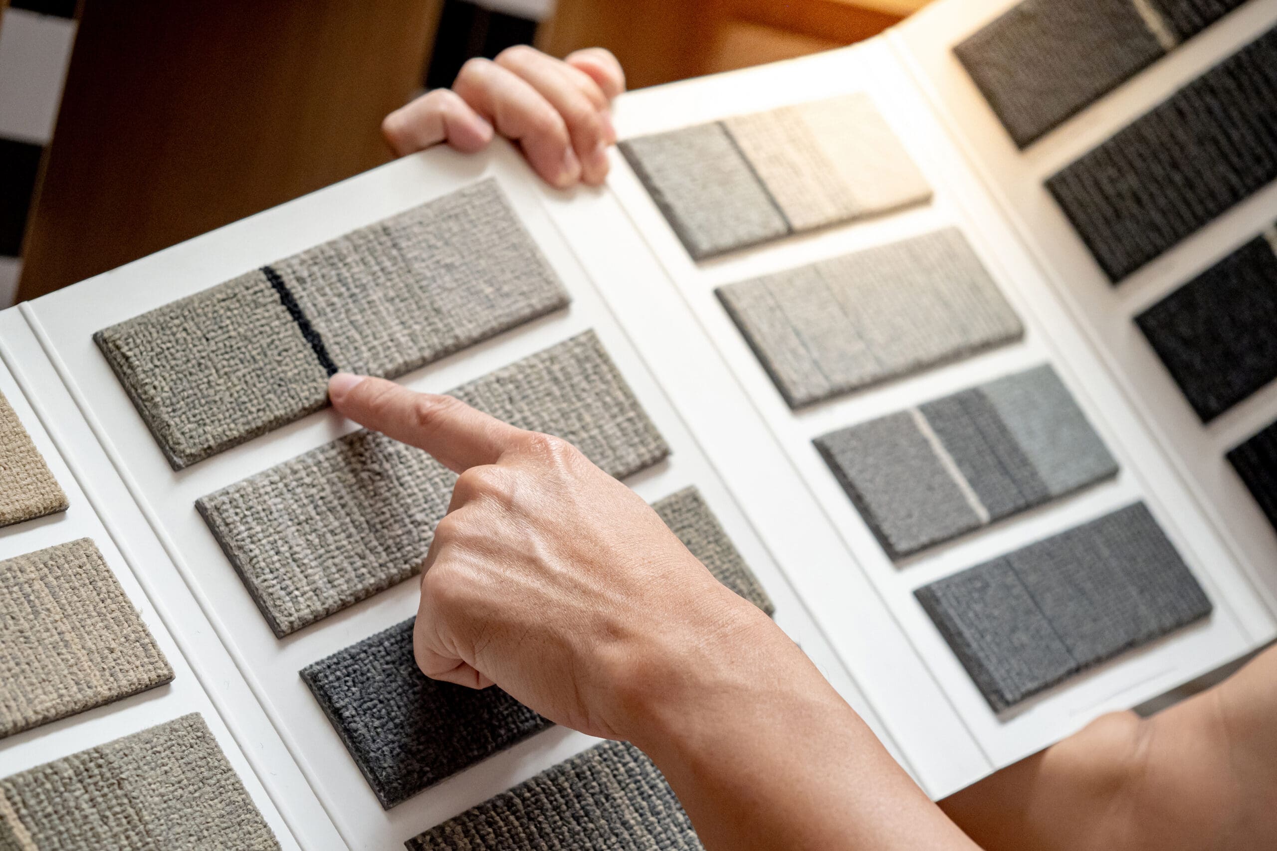 A person points at a sample in a display book of carpet swatches, exploring various shades and textures—an inspiring moment for anyone considering Flooring Installation or refreshing their space with new Decorating ideas.